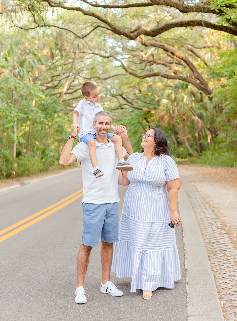 Father holding son on his shoulders, mother standing next to them. Son and mother smile at each other while father smiles at the camera. Taken at Philippe Park, FL during a family photo session showcasing outfit inspiration