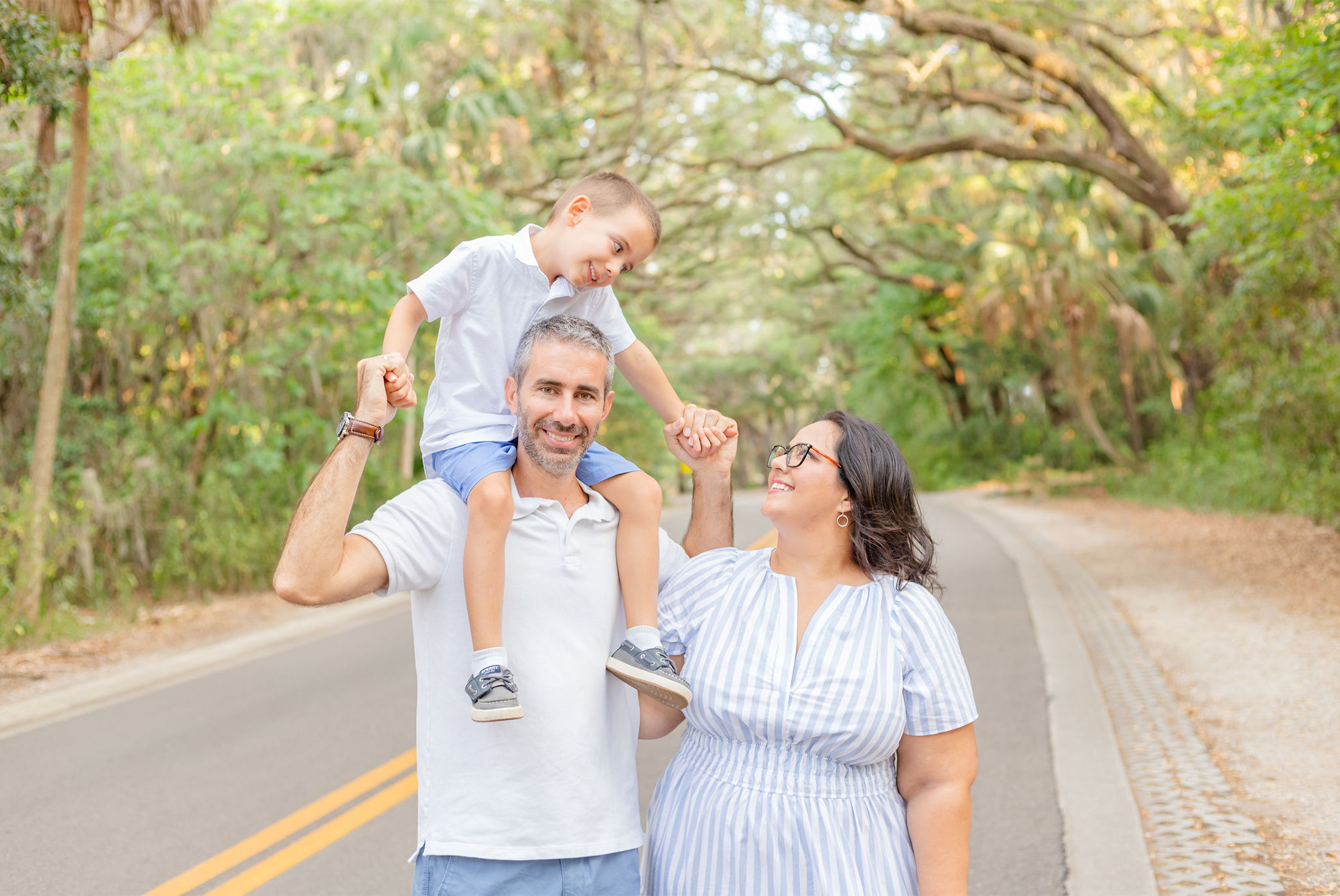 Father holding son on his shoulders, mother standing next to them. Son and mother smile at each other while father smiles at the camera. Taken at Philippe Park, FL during a family photo session showcasing outfit inspiration