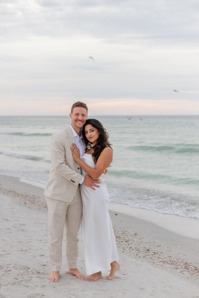 Bride Siddhi in a white satin dress and groom Zach in a beige suit embracing the beach.