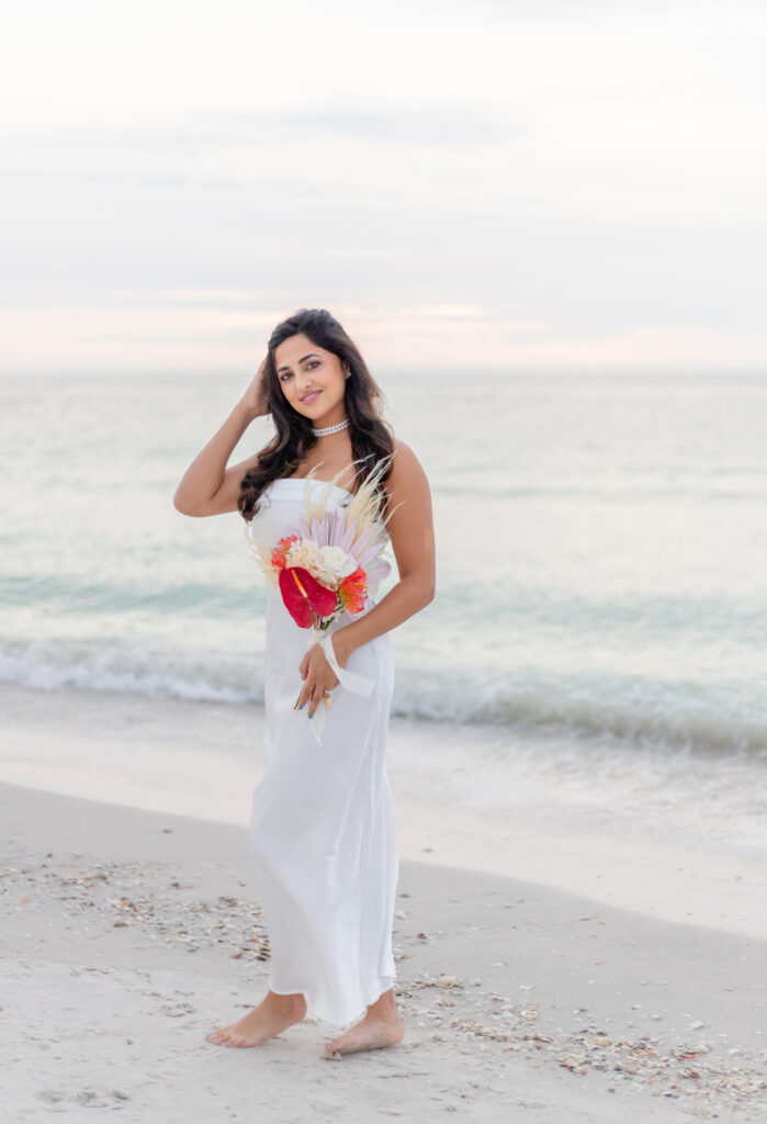 Bride Siddhi in a white satin dress standing on the beach at Saint Pete with ocean waves in the background.