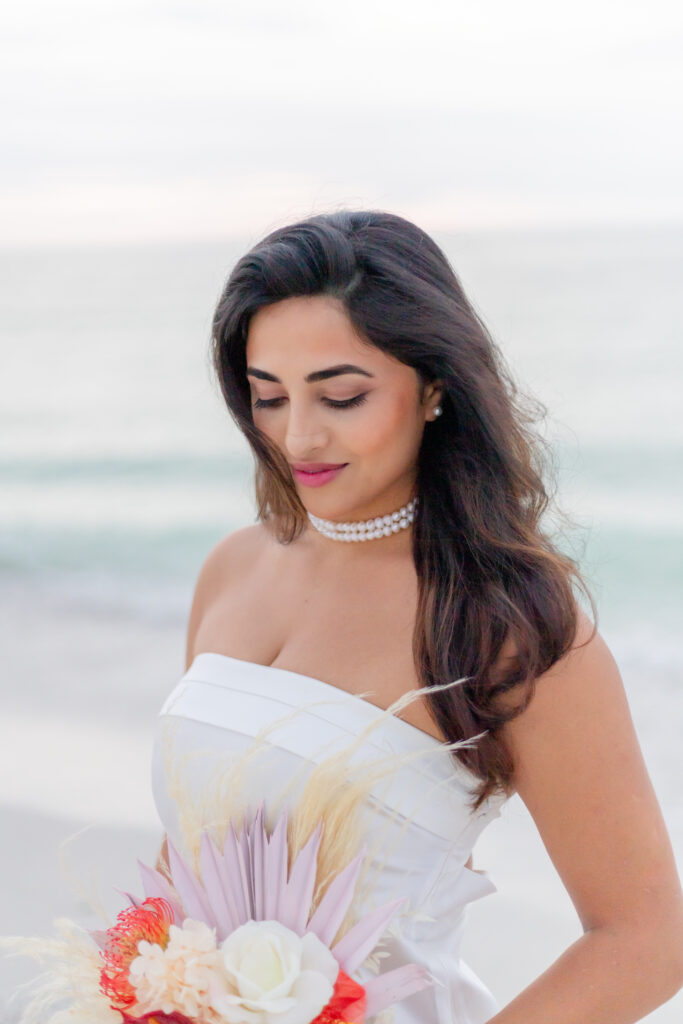 Bride Siddhi in a white satin dress standing on the beach at Saint Pete with ocean waves in the background.