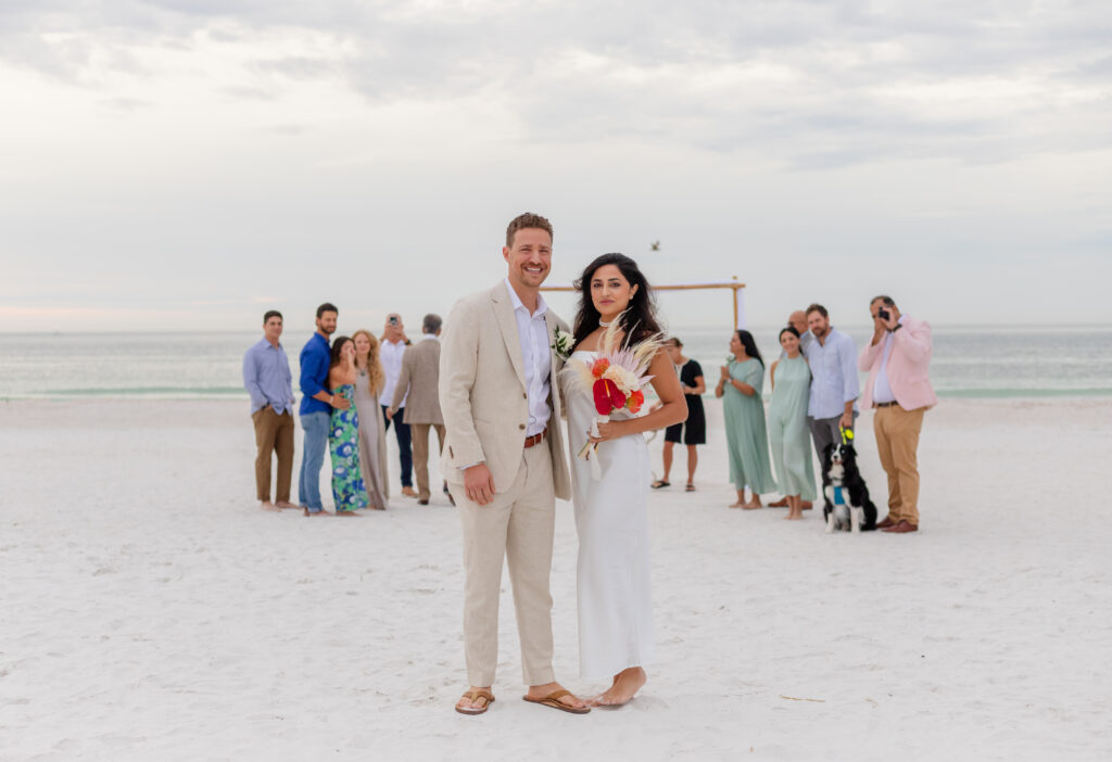 Bride Siddhi in a white satin dress and groom Zach in a beige suit embracing the beach against the sunset, with guests in pastel colors.