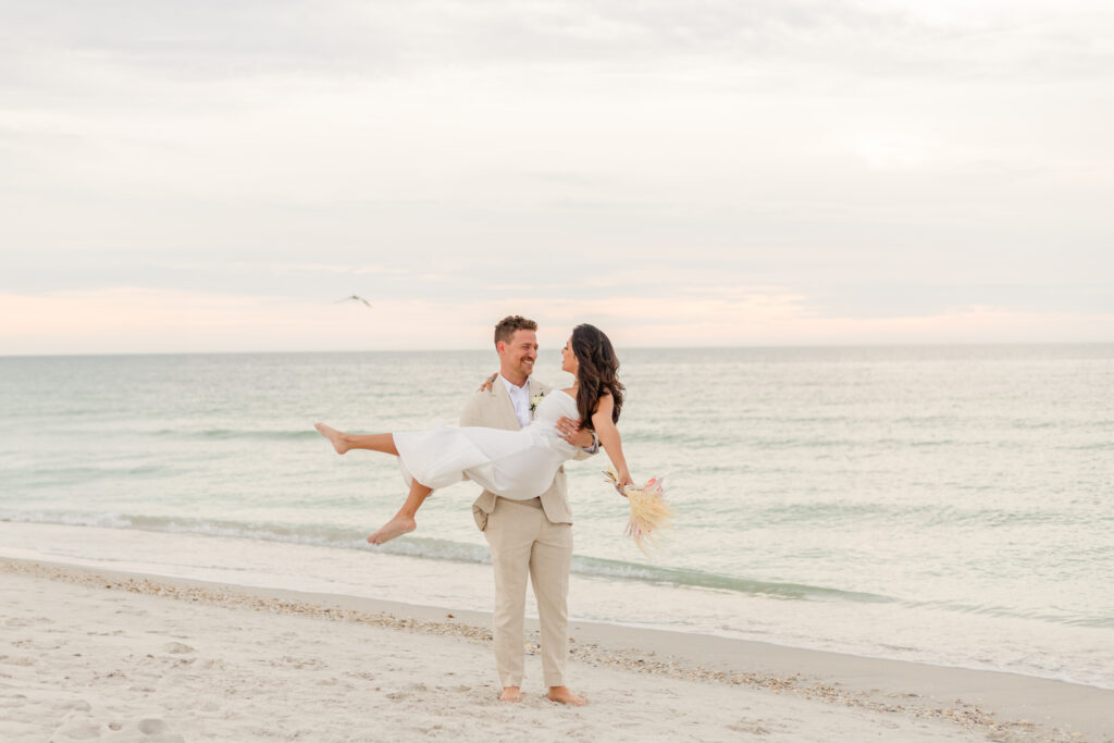 Groom Zach holding bride Siddhi in his arms, both smiling happily at their beach wedding.